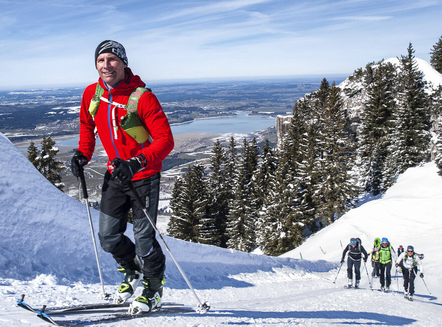 Das Bild zeigt einen Skitourengeher mit roter Jacke und schwarzer Mütze der bei schönem Wetter entlang einer präparierten Strecke bergauf durch den Schnee läuft. Er lächelt. Im Hintergrund sind weitere Skitourengeher zu erkennen.
