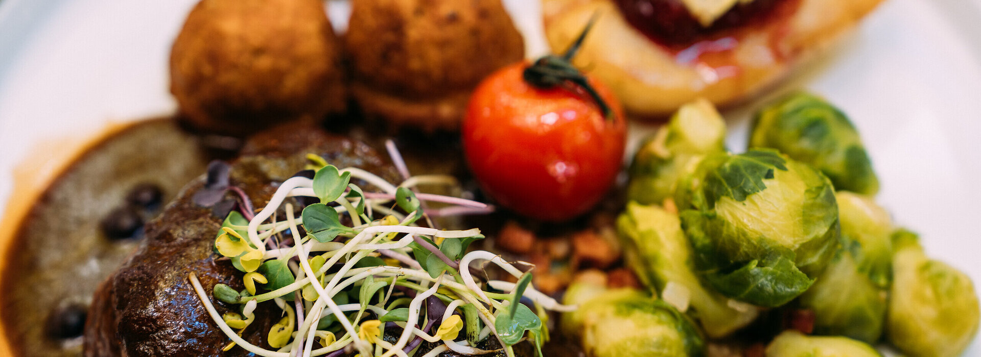 Ein appetitliches Gericht mit einem Fleischbraten, Rosenkohl, Kartoffelklößen und einer Tomate wird auf einem Teller präsentiert.
