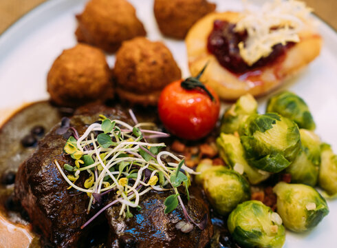 Ein appetitliches Gericht mit einem Fleischbraten, Rosenkohl, Kartoffelklößen und einer Tomate wird auf einem Teller präsentiert.