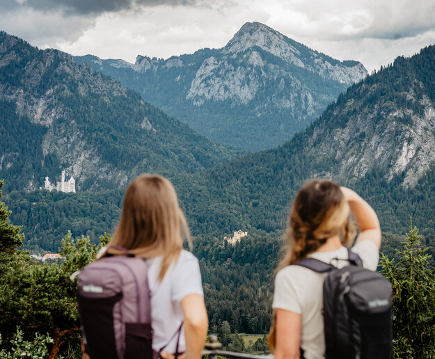 Das Bild zeigt zwei Frauen mit Rucksäcken von hinten, die in die Ferne schauen. Im Hintergrund erheben sich dunkle Wälder, die Königsschlösser sowie die Bergwelt der Ammergauer Alpen.