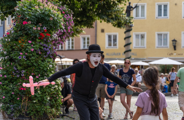 Das Bild zeigt einen Pantomimen, der ein Luftballontier in der rechten Hand hält. Er ist schwarz gekleidet, trägt einen schwarzen Hut und ist weiß geschminkt. Er steht in einer Fußgängerzone. Mehrere Personen sind zu sehen. Im Hintergrund sieht man bunte Hausfassaden. Die Sonne scheint. 