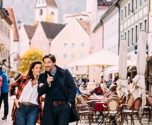 Ein Paar strahlt und läuft lachend auf einem Kopfsteinpflasterweg durch die malerische Füssener Altstadt mit traditionellen Häusern und Cafés.
