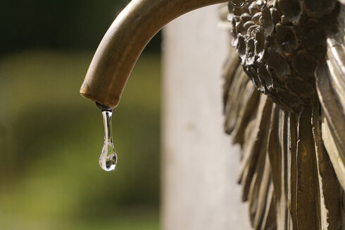 Zu sehen ist die Nahaufnahme eines messingfarbenen Wasserhahns, aus dem ein einzelner Wassertropfen herab perlt. Der Wasserhahn kommt aus einer messingfarbenen Arnikablüte, die an einem aufrecht stehenden Stein angebracht ist.