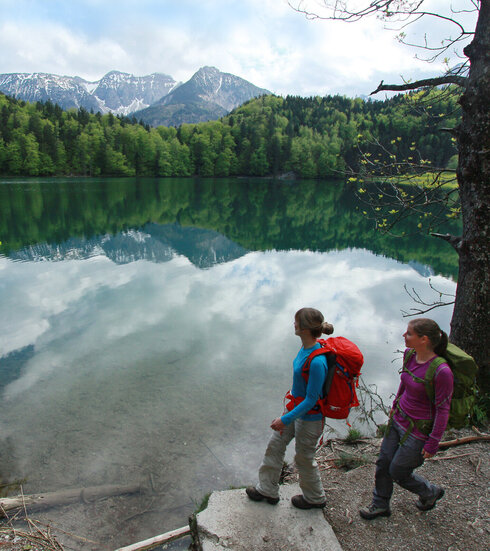 Auf dem Bild sieht man zwie Frauen mit Wanderkleidung und Wanderrucksäcken die an einem Seeufer laufen. Am Seeufer sind Bäumde und im Hintergrund in der Ferne schneebedkeckte Berge.