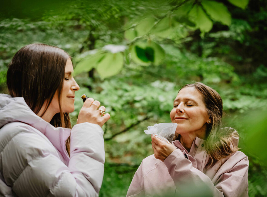 Das Bild zeigt zwei Frauen, die im Wald an Kräutern und ätherischen Ölen riechen