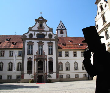 Das Foto zeigt den Klosterhof des ehemaligen Benediktinerklosters St. Mang. Der Blick fällt auf den Eingang des Museums der Stadt Füssen. Der Boden des Hofs ist mit Kopfsteinpflaster gepflastert. Die Fassade des Gebäudes ist mit Illusionsmalerei verziert. Vor dem Eingang des Museums liegt ein roter Teppich auf dem Boden. Der Himmel ist blau. Die Sonne scheint.