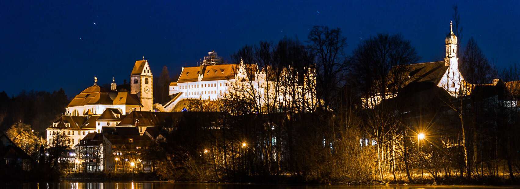 Das Bild ist eine Nachtaufnahme des Kloster St. Mang in Füssen, einem schlossartigen Gebäude. Die ehemalige Benediktiner Kloster spiegelt sich im Wasser wider, was ein Spiegelbild schafft.