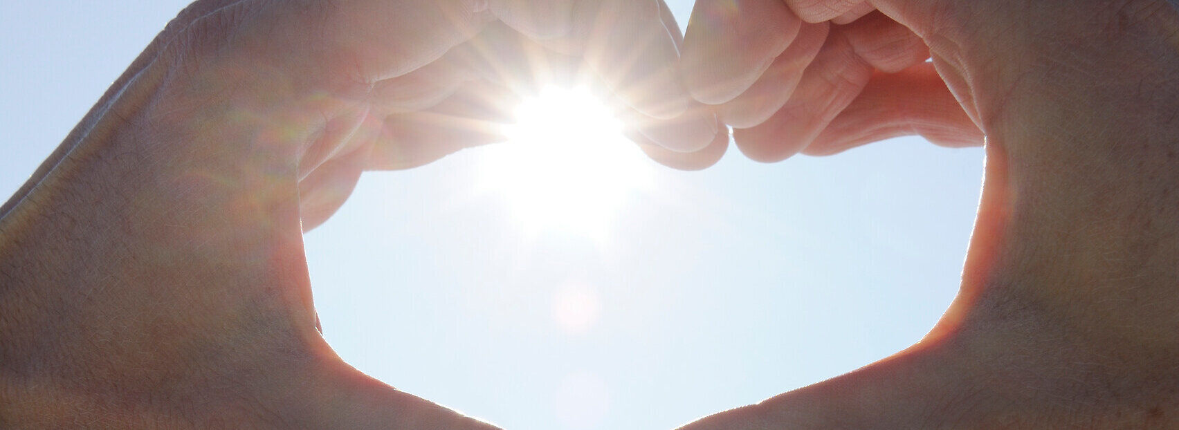 Das Bild zeigt zwei Hände, die ein Herz formen. Durch die Hände strahlt die Wintersonne vor dem blauen, klaren Himmel.
