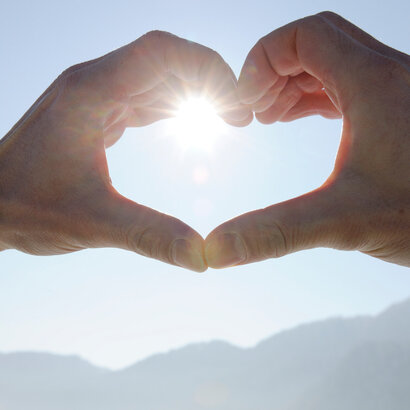 Das Bild zeigt zwei Hände, die ein Herz formen. Durch die Hände strahlt die Wintersonne vor dem blauen, klaren Himmel.