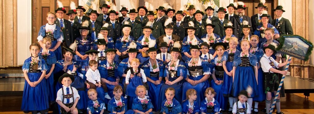 Eine große Gruppe von Menschen in traditioneller bayerischer Tracht, darunter viele Kinder, posiert in einem festlichen Raum. Sie tragen passende Hüte und Zierblumen. Im Hintergrund sind dekorative Wandmalereien und Fahnen zu sehen, die zur festlichen Atmosphäre beitragen.