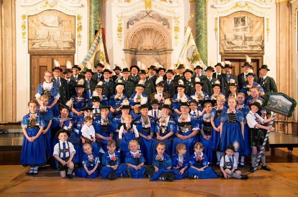 Eine große Gruppe von Menschen in traditioneller bayerischer Tracht, darunter viele Kinder, posiert in einem festlichen Raum. Sie tragen passende Hüte und Zierblumen. Im Hintergrund sind dekorative Wandmalereien und Fahnen zu sehen, die zur festlichen Atmosphäre beitragen.