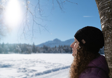 Eine Frau mit dunklen, langen Locken und schwarzer Strickmütze lehnt sich mit geschlossenen Augen gegen einen Baumstamm. Die Sonne strahlt ihr ins Gesicht und im Hintergrund ist eine Winterlandschaft mit schneebedeckten Bergen zu sehen.
