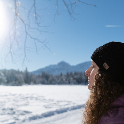 Eine Frau mit dunklen, langen Locken und schwarzer Strickmütze lehnt sich mit geschlossenen Augen gegen einen Baumstamm. Die Sonne strahlt ihr ins Gesicht und im Hintergrund ist eine Winterlandschaft mit schneebedeckten Bergen zu sehen.