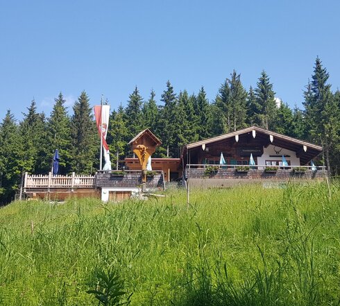 Das Bild zeigt eine große Holzhütte auf einem grünen Hügel mit einer großen Holzterrasse. Eine Fahne an einem großen Fahnenmast weht im Wind. Im Hintergrund ist ein dunkler Nadelwald.