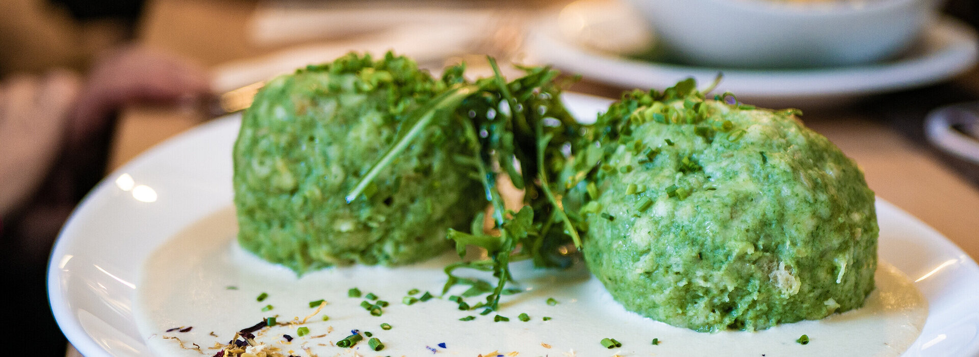 Auf einem weißen Teller liegen zwei Spinatknödel, die mit Kräutern und essbaren Blüten garniert sind. Das Gericht wird auf einem Holztisch serviert, und im Hintergrund sind weitere Teller zu sehen.
