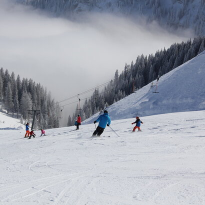 Das Bild zeigt eine abfallende Skipiste auf der mehrere Personen, auch Kinder Skifahren. Neben der Piste läuft der Schlepplift. Im Tal liegt eine dicke Nebelschicht. Die Landschaft ringsum ist tief verschneit. Die Sonne scheint.  