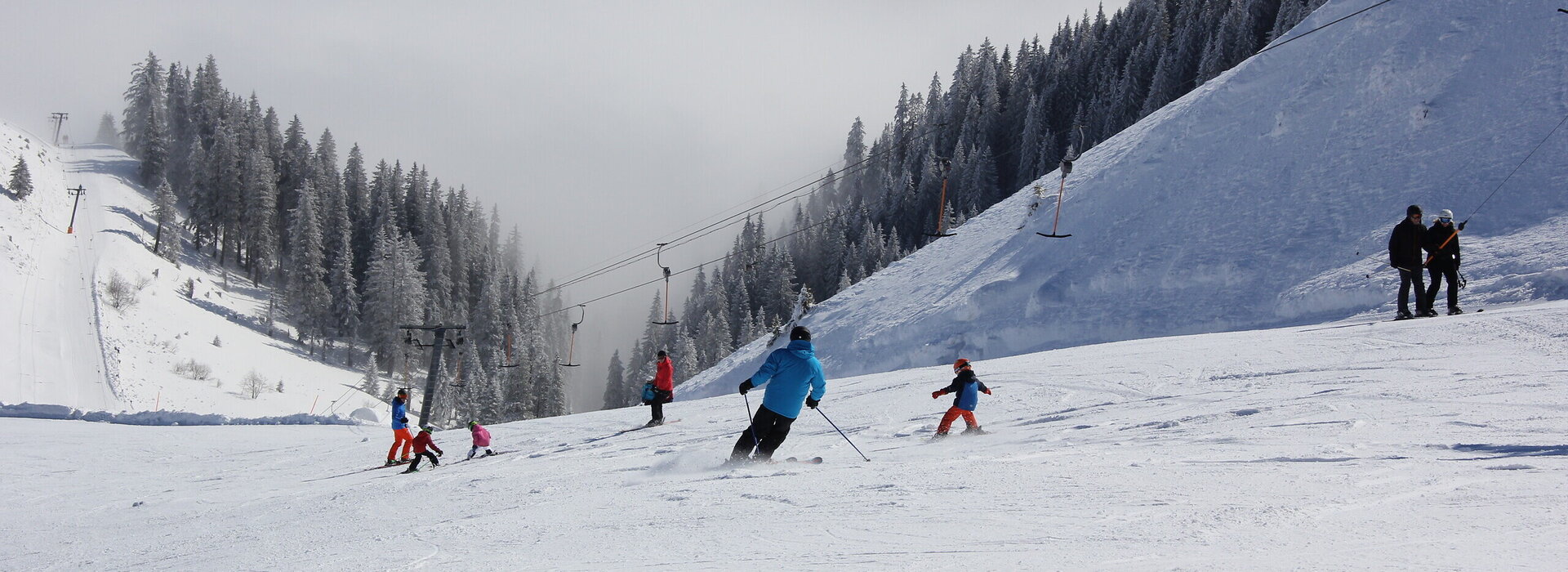 Das Bild zeigt eine abfallende Skipiste auf der mehrere Personen, auch Kinder Skifahren. Neben der Piste läuft der Schlepplift. Im Tal liegt eine dicke Nebelschicht. Die Landschaft ringsum ist tief verschneit. Die Sonne scheint.  