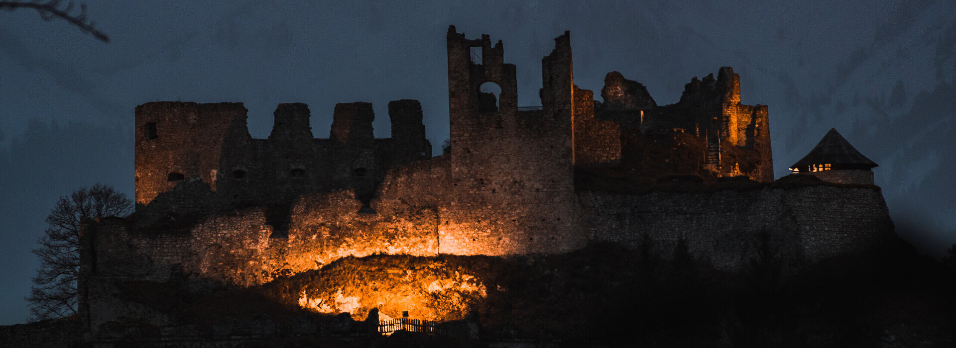 Das Bild zeigt eine beleuchtete Burgruine in der Abenddämmerung. Im Hintergrund erheben sich schneebedeckte Berge.  