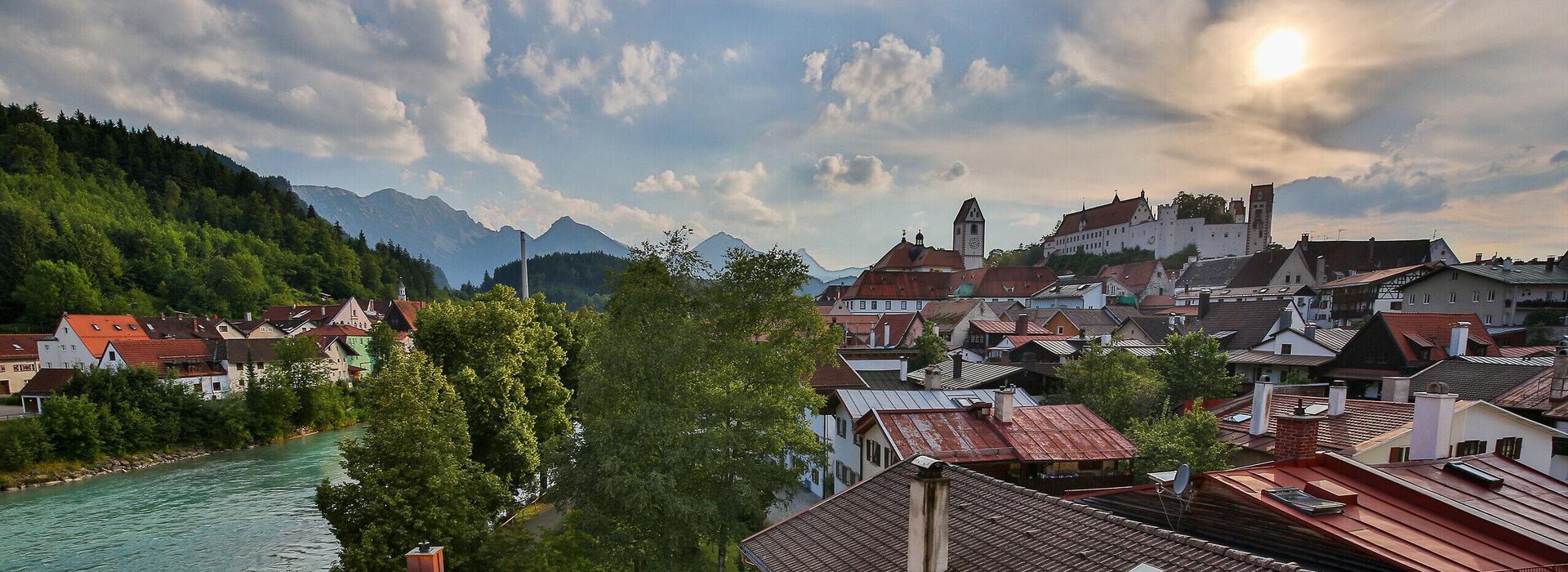 Die malerische Füssener Altstadt mit roten Dächern erstreckt sich am Ufer des Leches. Im Hintergrund rücken majestätische Berge in den Himmel.