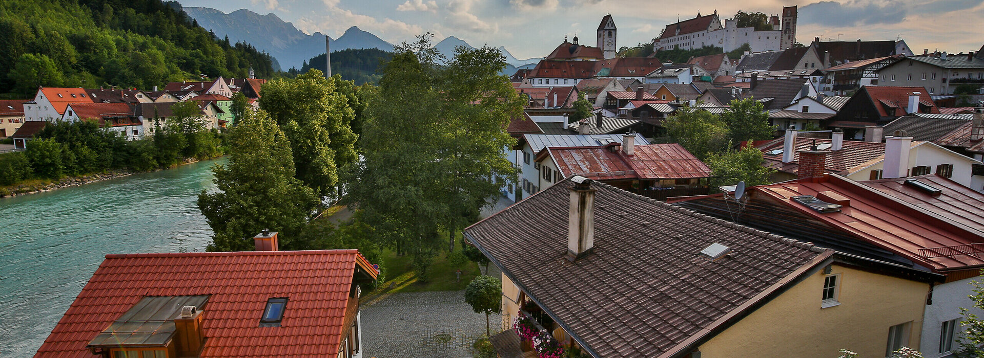 Die malerische Füssener Altstadt mit roten Dächern erstreckt sich am Ufer des Leches. Im Hintergrund rücken majestätische Berge in den Himmel.