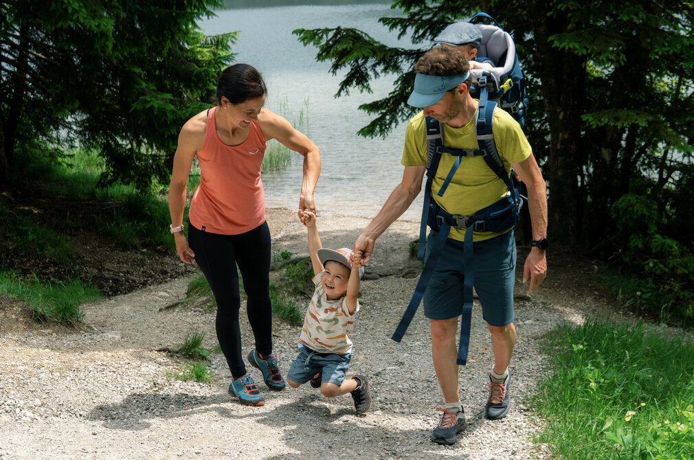 Eine Frau und ein Mann gehen gemeinsam mit einem fröhlichen Kind an einem Weg entlang, der zu einem See führt. Die Frau hält das Kind an der Hand, während der Mann ein Kind auf dem Rücken trägt. Um sie herum sind Bäume und grünes Gras.