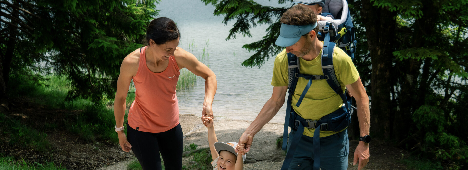 Eine Frau und ein Mann gehen gemeinsam mit einem fröhlichen Kind an einem Weg entlang, der zu einem See führt. Die Frau hält das Kind an der Hand, während der Mann ein Kind auf dem Rücken trägt. Um sie herum sind Bäume und grünes Gras.
