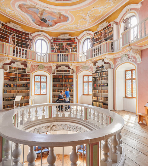 Die Bibliothek des Hohen Schlosses in Füssen mit hohen Regalen und einem beeindruckenden Rundbogen. Das Bild zeigt einen Raum voller Wissen und Geschichte.