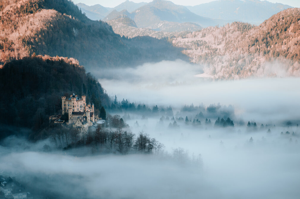 Schloss Hohenschwangau mit Türmen auf bewaldetem Hügel, umgeben von dichtem Nebel im Tal und Bergen im Hintergrund.
