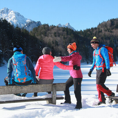 Das Bild zeigt vier bunt gekleidete Personen in einer winterlichen Landschaft. Zwei Personen sitzen auf der Lehne einer Bank, während die anderen beiden Personen begrüßend auf sie zukommen. Im Hintergrund sind ein gefrorener See und schneebedeckte Berge zu erkennen.