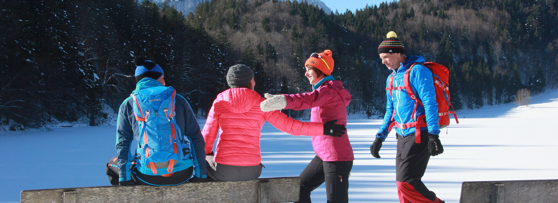 Das Bild zeigt vier bunt gekleidete Personen in einer winterlichen Landschaft. Zwei Personen sitzen auf der Lehne einer Bank, während die anderen beiden Personen begrüßend auf sie zukommen. Im Hintergrund sind ein gefrorener See und schneebedeckte Berge zu erkennen.