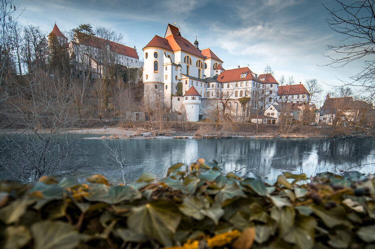 Schloss und Kloster mit roten Dächern und Türmen am Ufer des Lech im Winter, Spiegelung im Wasser, Vordergrund durch unscharfen Efeu verdeckt.