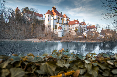 Schloss und Kloster mit roten Dächern und Türmen am Ufer des Lech im Winter, Spiegelung im Wasser, Vordergrund durch unscharfen Efeu verdeckt.