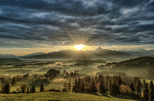 Das Bild zeigt eine atemberaubende Landschaft, unter einem dramatischen Himmel. Der Himmel ist voller dunkler Wolken, die eine Kulisse für die Szene schaffen. Die Sonne scheint hell durch die Wolken und gibt der Landschaft darunter ein warmes Glühen. Die Berge in der Ferne sind majestätisch, mit ihren Spitzen streben sie zum Himmel. Die Vordergrund ist voller grüner Bäume, die einen Farbtupfer in die ansonsten monochrome Szene bringen. Die Gesamtzusammenstellung des Bildes erzeugt ein Gefühl von Ehrfurcht und Bewunderung für die Schönheit der Natur.