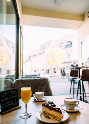 Ein Glas Orangensaft, ein Stück Kuchen und eine Tasse Kaffee stehen auf einem Holztisch in einem Café mit Blick in die Füssener Altstadt.