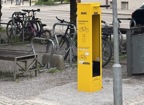 Ein Straßenschild zeigt verschiedene Richtungen mit Entfernungsangaben zu Roßhaupten, Forggensee, Hohenschwangau und Schwangau. In der Nähe steht eine gelbe Radservice-Station und Fahrräder sind an einem Gestell geparkt. Im Hintergrund sind Bäume und ein Gebäude sichtbar.