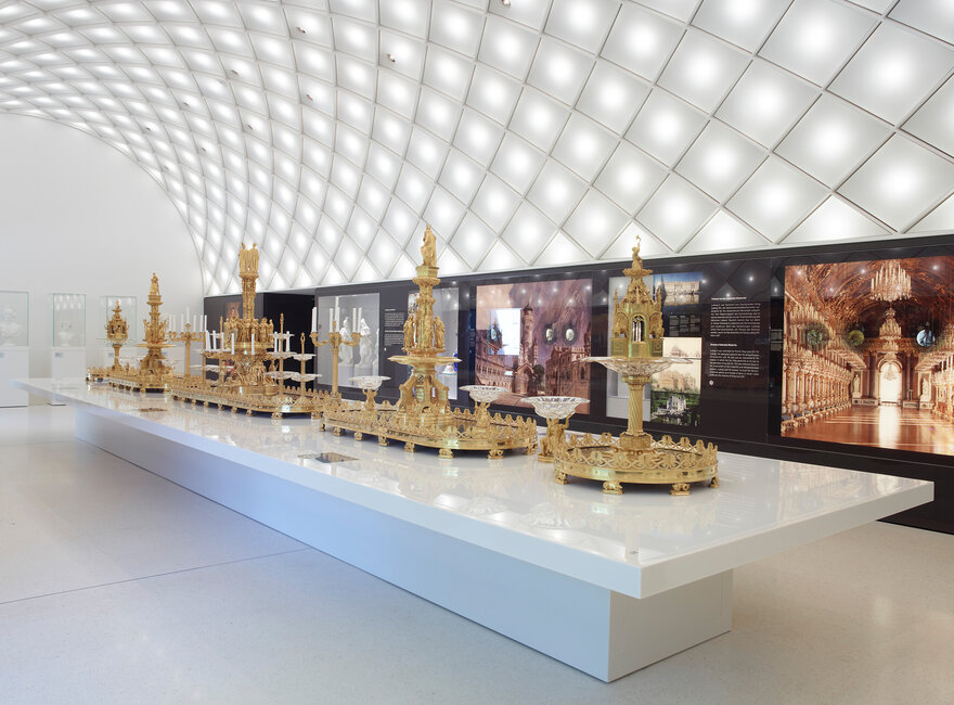 Goldene Tafelaufsätze auf weißem Ausstellungstisch unter gewölbter Decke mit Rautenstruktur im Museum der bayerischen Könige.