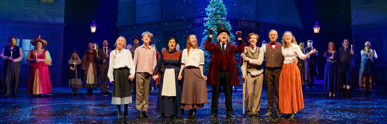 Musicalbühne mit blauer Beleuchtung und Kulisse von Häusern sowie einem Weihnachtsbaum. Viele Schauspieler in Kostümen des 19. Jahrhunderts stehen darauf.