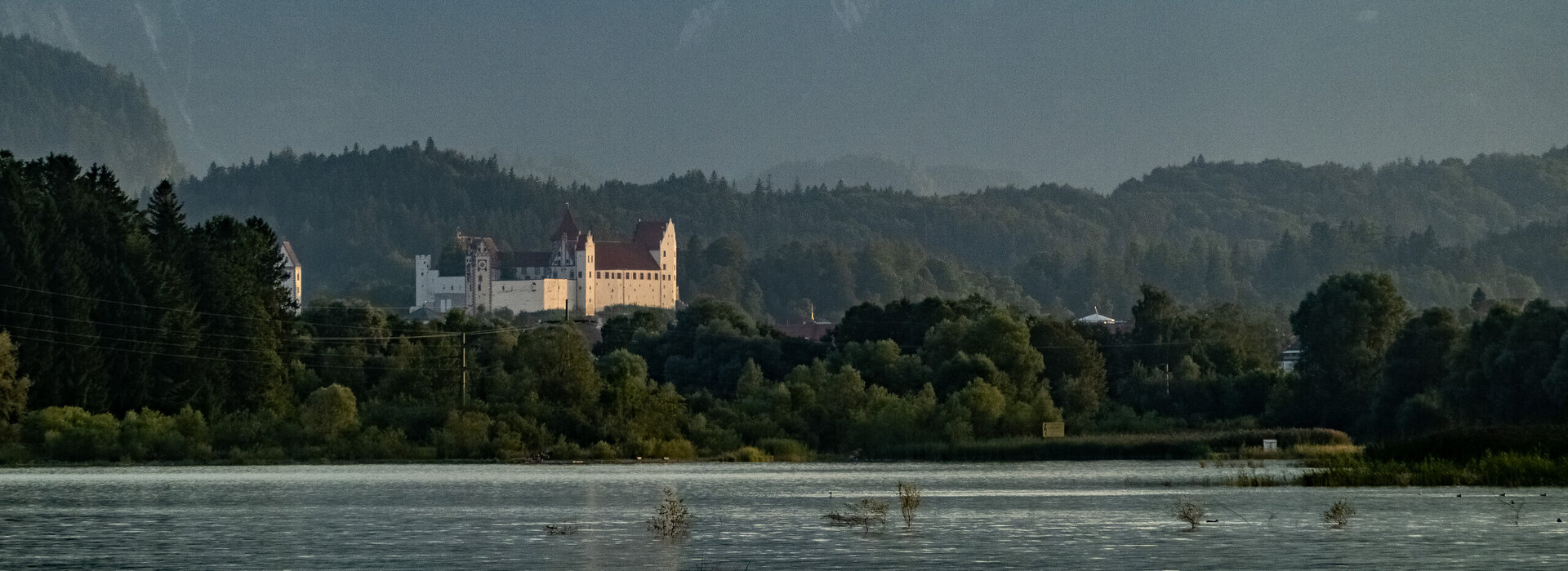 Das Bild zeigt eine friedliche Landschaft, die von einem majestätischen Schloss dominiert wird. Es handelt sich hier um das Hohe Schloss bei Füssen, das mit seinen weißen Wänden und dem roten Dach vor dem Bergpanorama steht. 