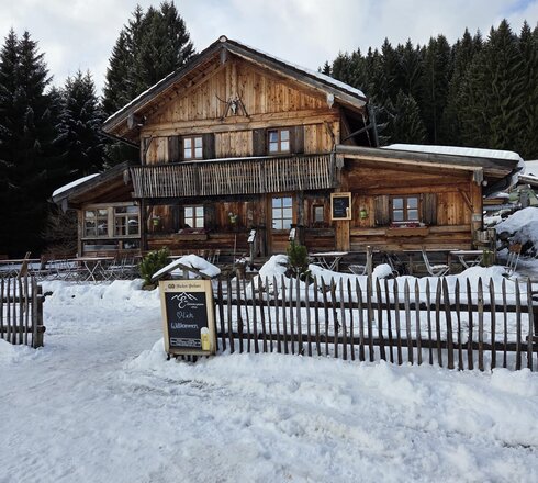 Das Bild zeigt eine schöne, große Holzhütte im Winter, die von einem Holzzaun umgeben ist. Im Hintergrund erhebt sich ein dunkler Nadelwald.