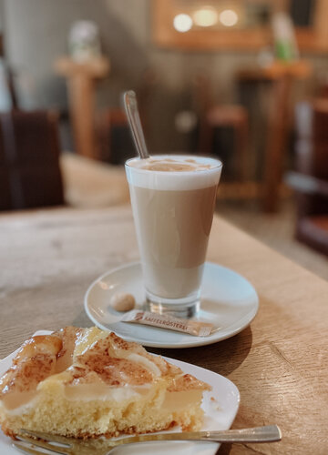 Dieses Bild zeigt einen Milchkaffee oder Latte, neben einem Stück Apfeltorte auf einem Untertasse. Die Szene vermittelt eine gemütliche Atmosphäre, die typisch für ein Café oder eine Bäckerei ist.