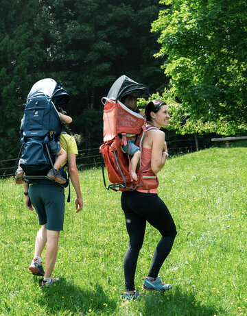 Zwei Erwachsene gehen auf einer grünen Wiese, jeder trägt einen kindgerechten Rucksack. Die Person links trägt einen dunkelblauen Rucksack, während die Person rechts einen roten Rucksack hat. Im Hintergrund sind Bäume zu sehen, die für eine natürliche Umgebung sorgen.