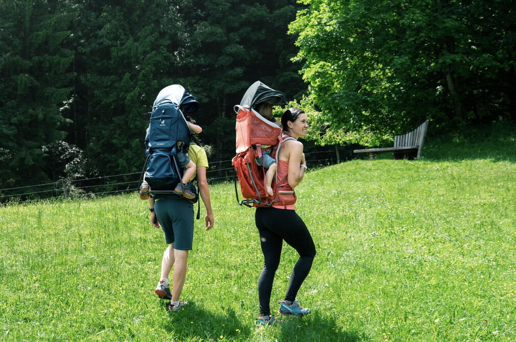 Zwei Erwachsene gehen auf einer grünen Wiese, jeder trägt einen kindgerechten Rucksack. Die Person links trägt einen dunkelblauen Rucksack, während die Person rechts einen roten Rucksack hat. Im Hintergrund sind Bäume zu sehen, die für eine natürliche Umgebung sorgen.