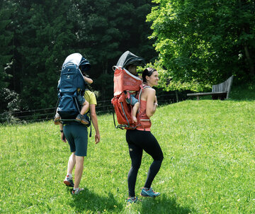 Zwei Erwachsene gehen auf einer grünen Wiese, jeder trägt einen kindgerechten Rucksack. Die Person links trägt einen dunkelblauen Rucksack, während die Person rechts einen roten Rucksack hat. Im Hintergrund sind Bäume zu sehen, die für eine natürliche Umgebung sorgen.