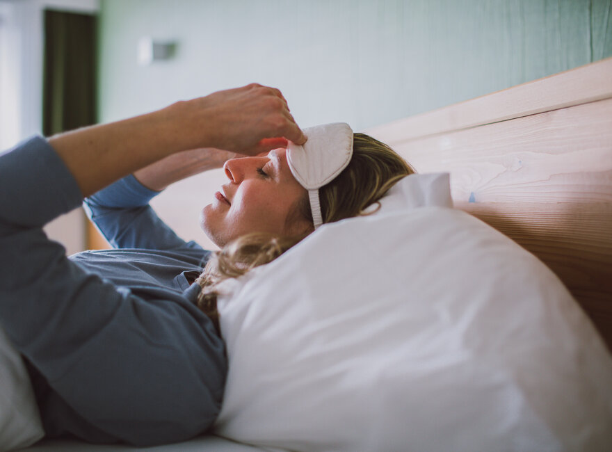Das Bild zeigt eine Frau mit einem blauen langen Oberteil. Sie liegt in einem Bett, das weiß bezogen ist und zieht sich gerade eine weiße Schlafmaske an.