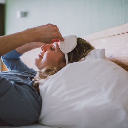 Das Bild zeigt eine Frau mit einem blauen langen Oberteil. Sie liegt in einem Bett, das weiß bezogen ist und zieht sich gerade eine weiße Schlafmaske an.