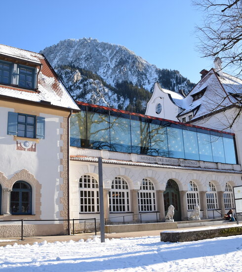 Traditionelles, weißes Gebäude mit moderner Glasbrücke, beschriftet "MUSEUM DER BAYERISCHEN KÖNIGE", umgeben von Schnee und Bergen.