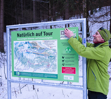 Das Bil zeigt einen Mann mit einer grünen Jacke und Mütze, der an einer großen Übersichtstafel zu Schutzgebieten steht. Im Hintergrund ist ein winterlicher Wald zu erkennen.