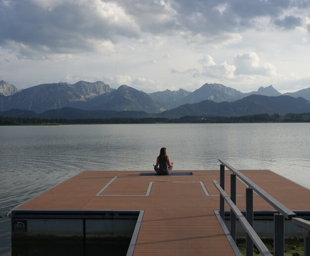 Eine Frau sitzt im Schneidersitz auf der Kneippinsel am Hopfensee mit Blick auf die Berge. 