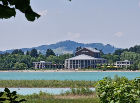 Ein Blick auf das Festspielhaus am Wasser, umgeben von grünen Bäumen und sanften Hügeln. Die ruhige, türkisfarbene Wasseroberfläche reflektiert den klaren Himmel. Im Vordergrund sind Schilf und Pflanzen zu sehen, die eine natürliche Umgebung schaffen.