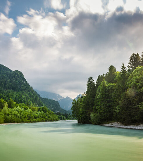 Das Bild zeigt eine friedliche Landschaft um den Fluss Lech, der durch einen grünen Wald fließt. Der Fluss, mit seinem leicht türkisenen Wasser, ist der zentrale Fokus des Bildes. Er windet sich durch den dichten Wald, erzeugt einen Sinn von Ruhe und Harmonie mit der Natur. Der Wald ist ein kräftiges Grün, gefüllt mit hohen Bäumen, die über den Fluss hineinwachsen. Der blaue Himmel darüber ist mit flauschigen weißen Wolken durchzogen, die zur Schönheit der Szene beitragen. Das Bild wird aus der Ferne aufgenommen, was einen umfassenden Blick auf den Fluss sowie den umliegenden Wald erlaubt. Die Perspektive des Bildes betont Tiefe, was die Weite des Waldes und des Flusses unterstreicht. Das Bild ist eine schöne Darstellung von Naturfrieden und der Harmonie zwischen dem Fluss und dem Wald.